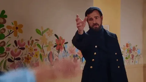 Animated gif of a bearded man in a navy gold-buttoned coat pointing, a floral mural behind him